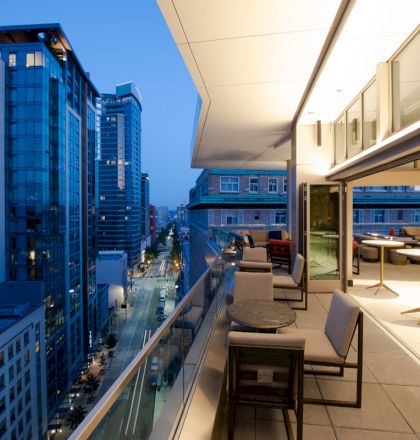 This image shows a modern outdoor patio on a high-rise building, with city buildings and a street visible in the background during evening time.