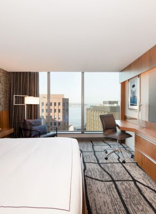 A modern hotel room with a large bed, flat-screen TV, desk, chair, and a window view of city buildings and the waterfront ending the sentence.