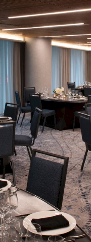 The image shows an elegantly set dining area with round tables and chairs, adorned with tableware, napkins, and floral centerpieces, in a well-lit room.