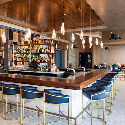 The image shows a modern bar with blue chairs, wooden countertops, hanging lights, and a fully stocked liquor display surrounded by open seating.