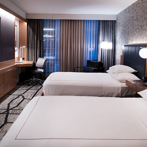 A modern hotel room features two neatly made beds, a flat-screen TV, a desk with a chair, and contemporary lighting and decor.