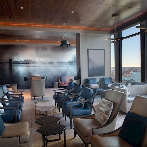 A modern lounge with plush seating, large windows showcasing a waterfront view and Ferris wheel, and ambient lighting and decor.
