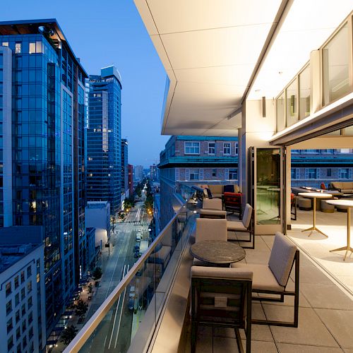 A modern cityscape during the evening featuring tall buildings, a street below, and a well-lit, furnished balcony with chairs and tables.