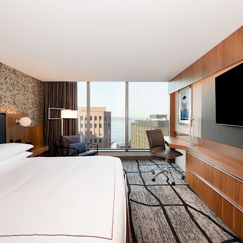 A modern hotel room with a large bed, wall-mounted TV, desk, chair, and a window showing a cityscape view, with contemporary decor.