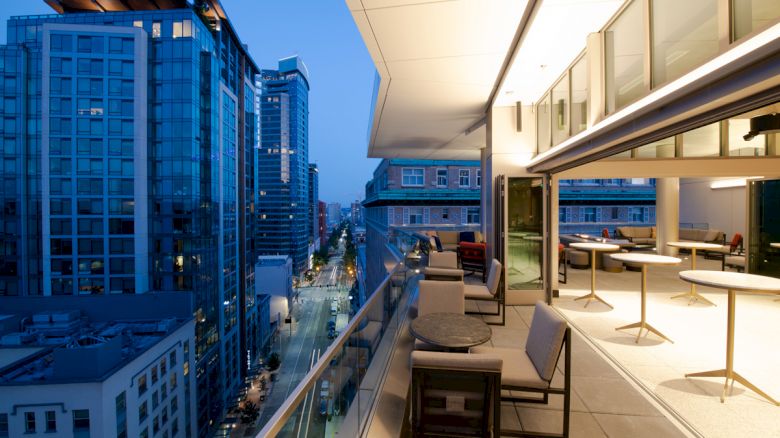 A modern balcony overlooks a city street with high-rise buildings. The balcony has seating and tables and is partially enclosed by glass doors.