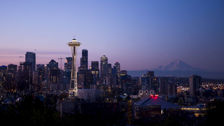 The image shows the Seattle skyline at dusk, featuring the Space Needle prominently, with Mount Rainier visible in the background.