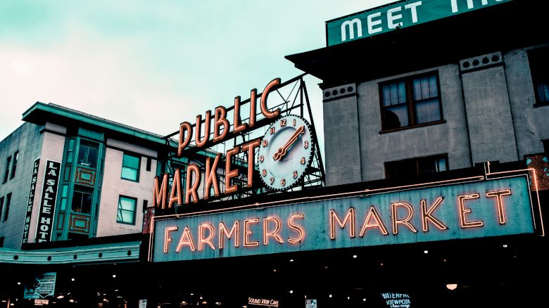 The image shows a "Public Market" sign with a clock and "Farmers Market" below, on the facade of a building. Another building's sign partially reads "MEET THE".