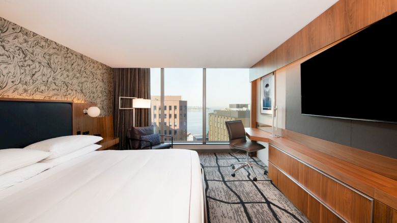 A modern hotel room with a large bed, a desk with a chair, a TV on the wall, and a window offering a cityscape view in the background.