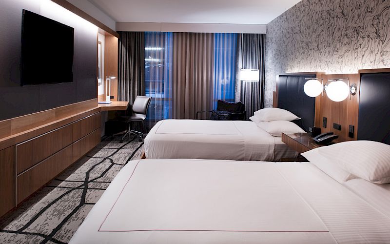 A modern hotel room with two beds, a wall-mounted TV, a workspace by the window, and stylish decor including patterned wallpaper and contemporary lighting.
