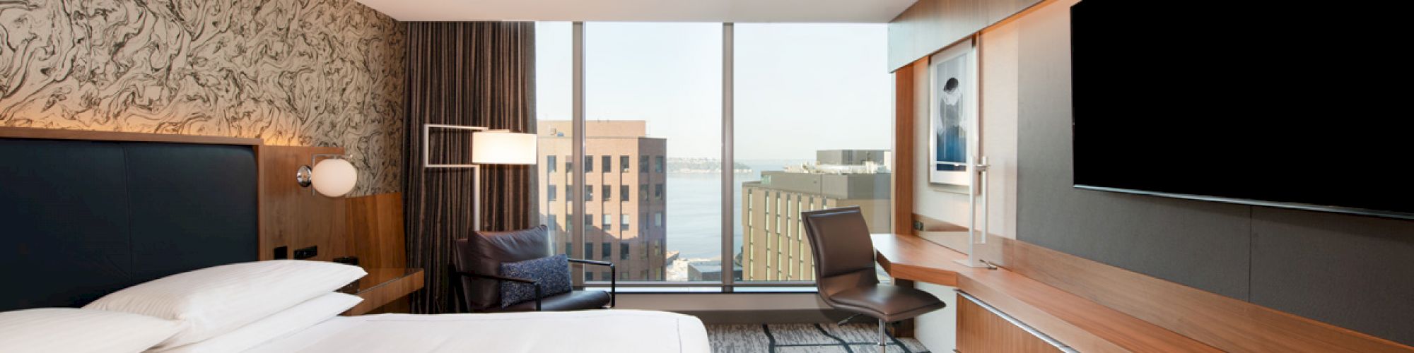 A modern hotel room features a large bed, a flat-screen TV, a work desk with a chair, and a view of city buildings through a large window.