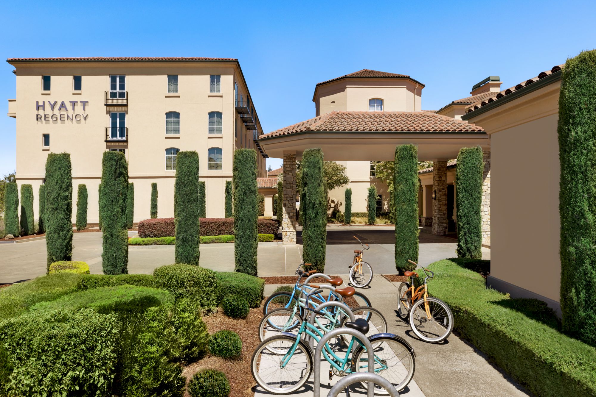 This image shows the exterior of a Hyatt Regency hotel with well-manicured landscaping and a bike rack with several bicycles parked.