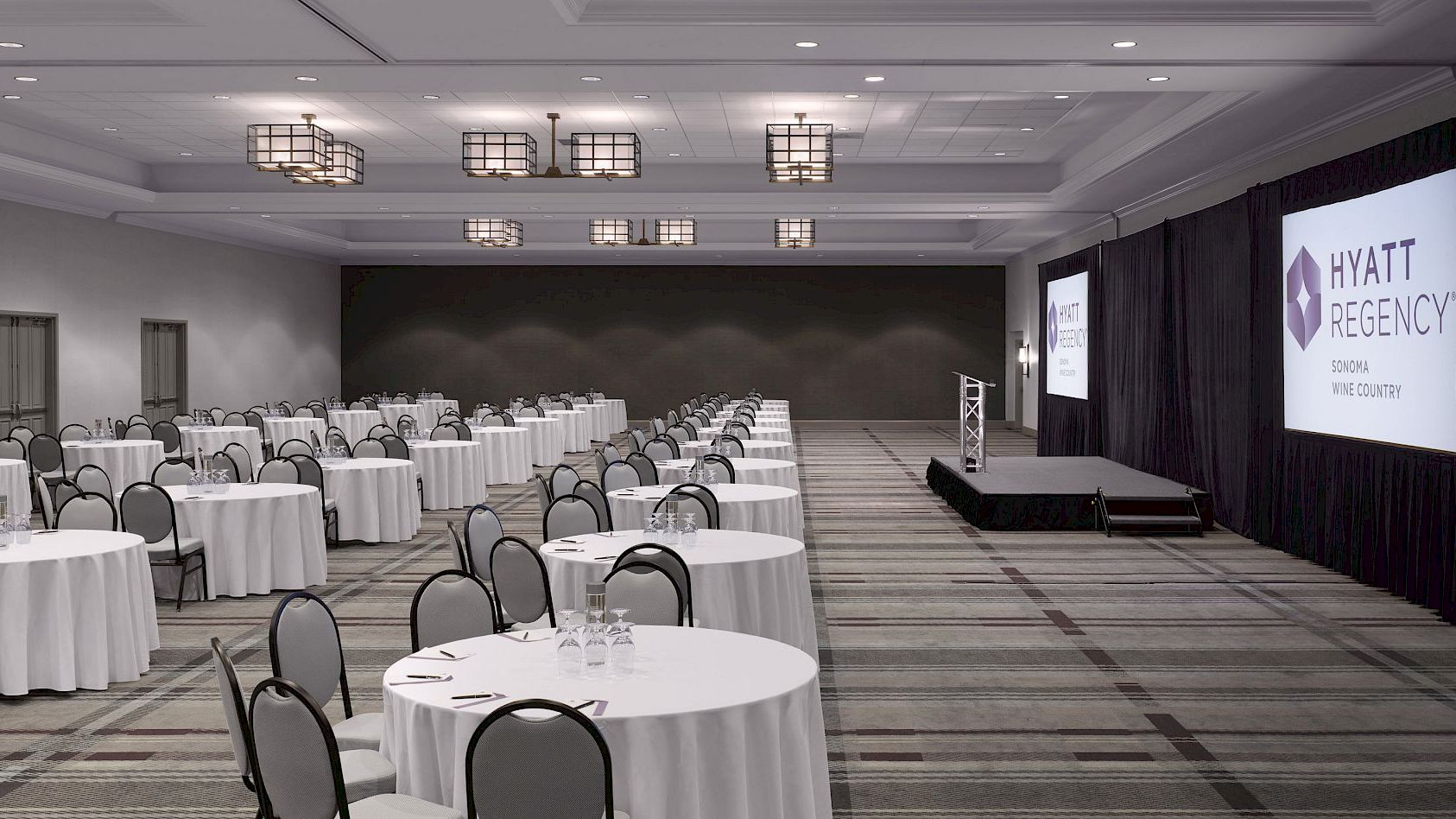A conference room with round tables, chairs, a stage, and screens displaying "HYATT REGENCY" on both sides of the stage.