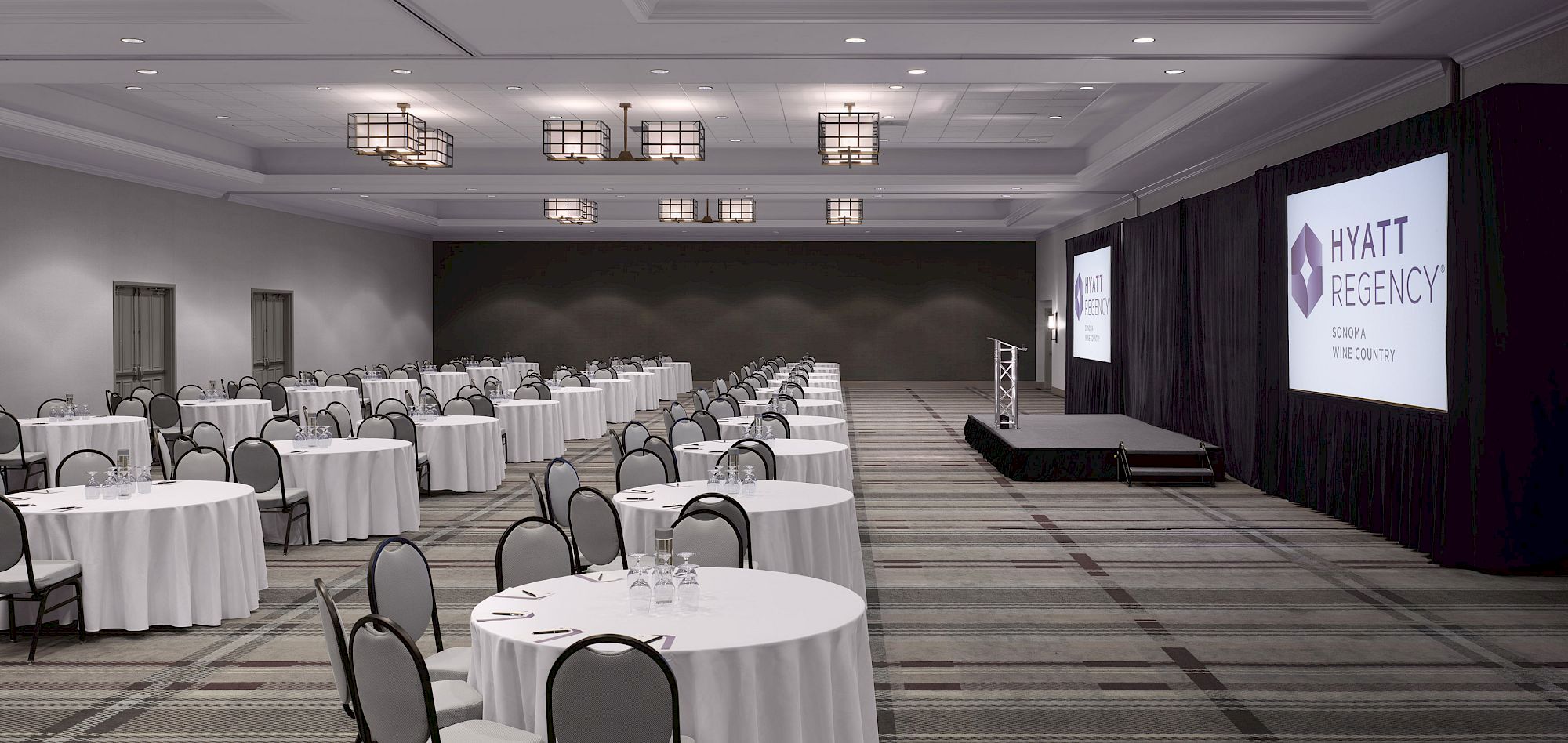 A conference room with round tables, chairs, a stage, and screens displaying 