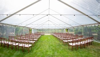 A large outdoor tent with a clear roof and a green grass floor, set up with rows of wooden chairs for a ceremony or event, likely a wedding.