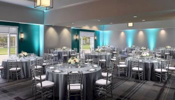 The image shows an elegantly decorated banquet hall with round tables set for an event, featuring floral centerpieces and neatly arranged place settings.