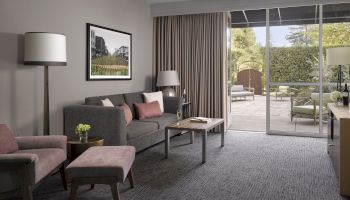 A modern living room with a sofa, armchair, floor lamp, coffee table, artwork, and patio doors leading to an outdoor area with seating.