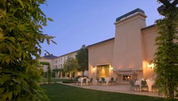 An elegant outdoor terrace with tables, chairs, and a large fireplace, surrounded by greenery, at a sophisticated building during dusk.