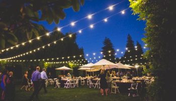 An outdoor evening event with string lights overhead, people mingling, and tables with white chairs and umbrellas are set up on a grassy area.