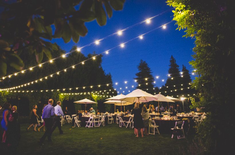 An outdoor evening event with string lights overhead, people mingling, and tables with white chairs and umbrellas are set up on a grassy area.